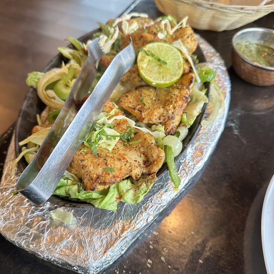 Sizzling Indian chicken tikka with lime, onions, and lettuce, served with tongs.